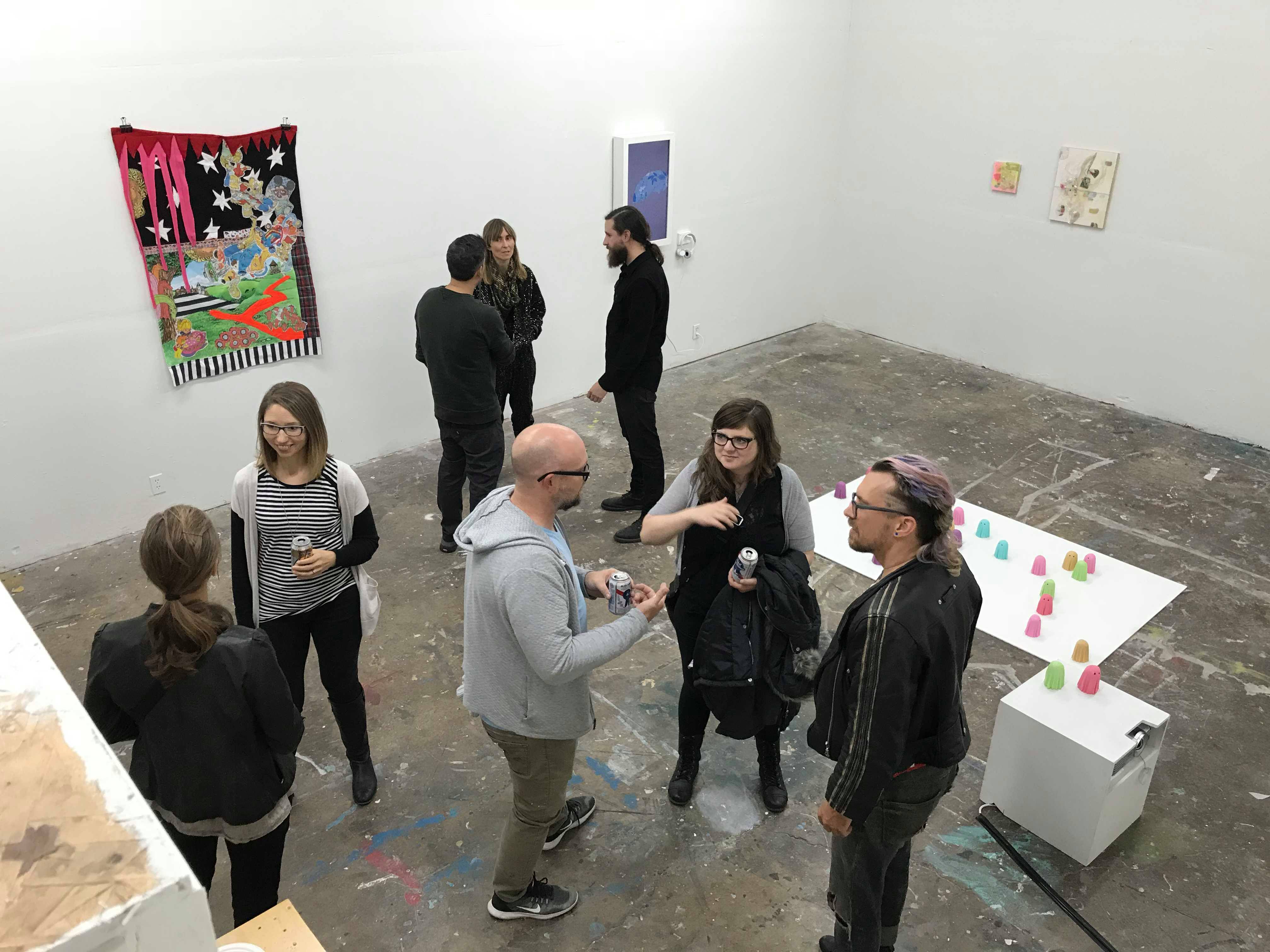 Overhead view of the white cube space of the art gallery Tropical Contemporary. There is art on the walls and on the floor. Small groups of people are standing together engaged in conversation.